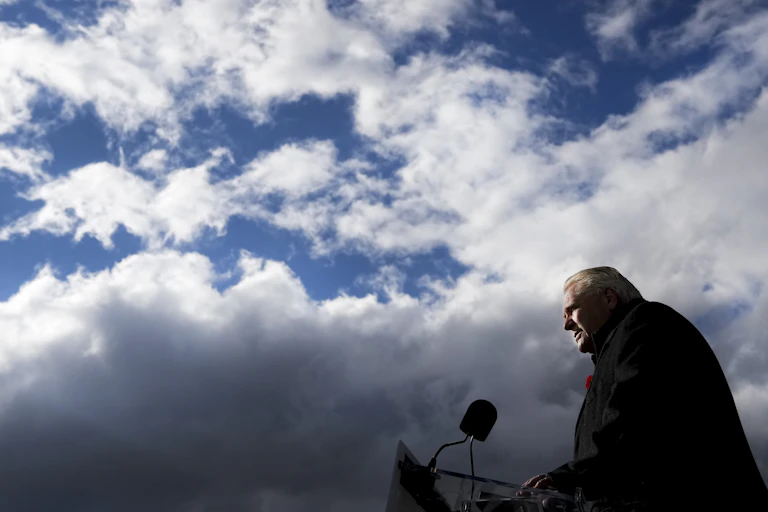 A man is seen from the side speaking into a microphone with blue sky and clouds above him.