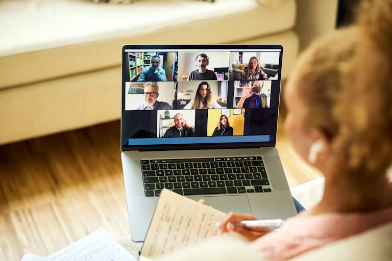 Woman taking notes as she stares at a computer screen during a videoconference with people staring back.