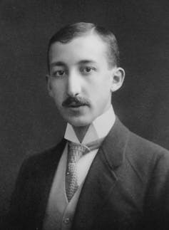 A black and white headshot of a young man with a mustache wearing a suit.