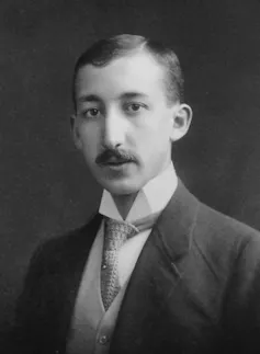 A black and white headshot of a young man with a mustache wearing a suit.