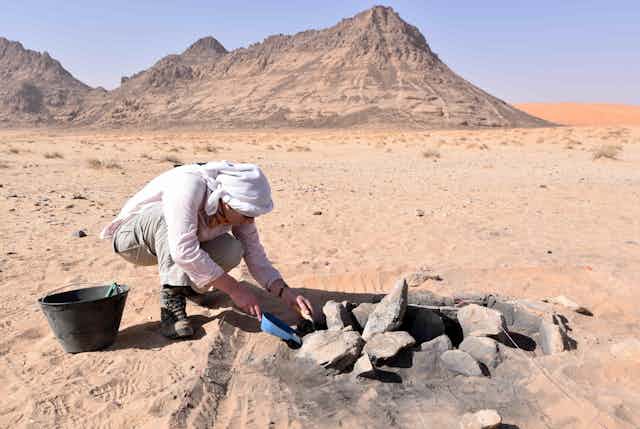 A photo of a person digging through rocks in a firepit in a desert, beneath a blue sky with rocky mountains in the background.