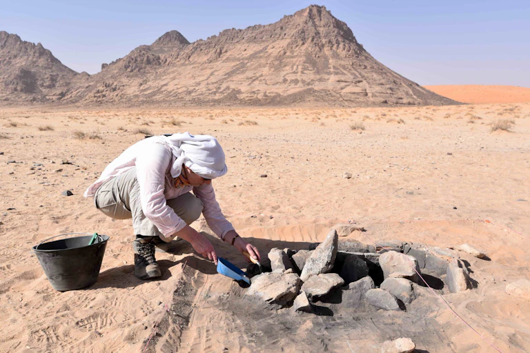 A photo of a person digging through rocks in a firepit in a desert, beneath a blue sky with rocky mountains in the background.