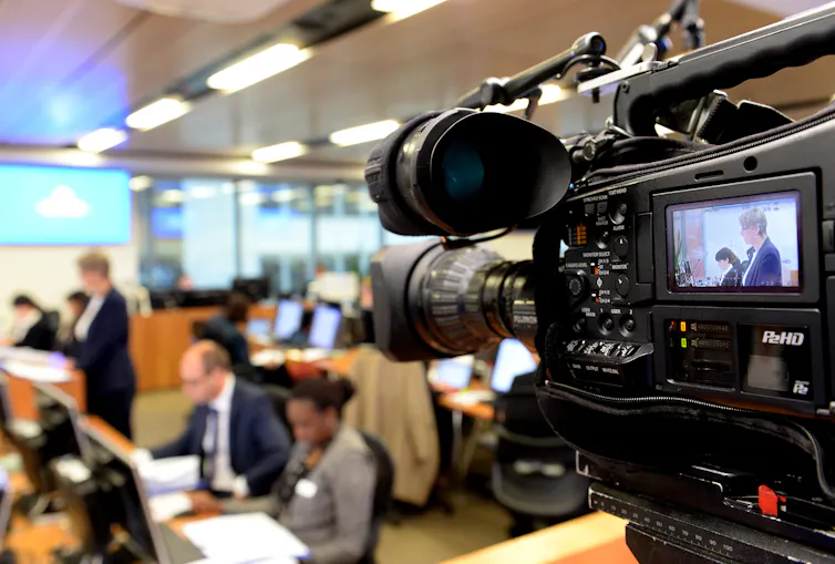 Video camera films a hearing room full of people.