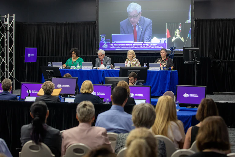 large screen shows man speaking in front of seated crowds, purple banners
