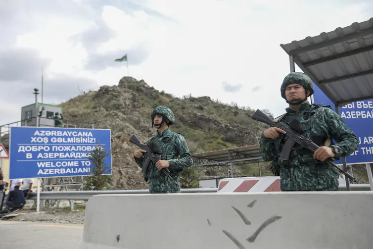 Men in uniform and holding guns stand by a concrete barrier.