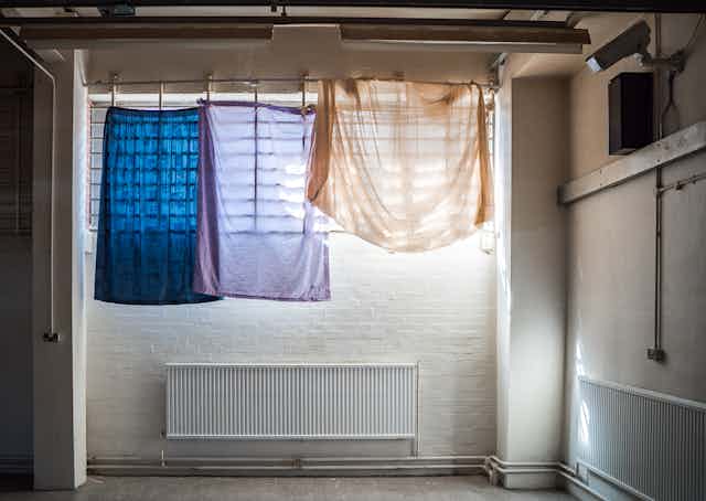 Coloured bedsheets hanging over a barred window inside a white, brick room with a CCTV camera on the wall