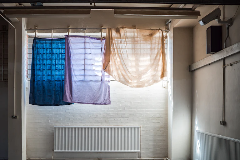Coloured bedsheets hanging over a barred window inside a white, brick room with a CCTV camera on the wall