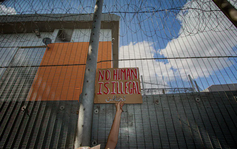 A single arm holding up a placard that reads 'no human being is illegal' against a barbed wire topped fence surrounding a building