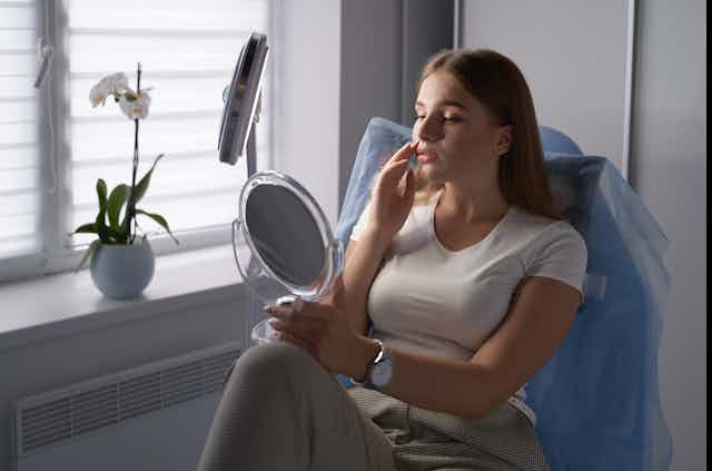 A young woman uses a mirror to scrutinise at her lips.