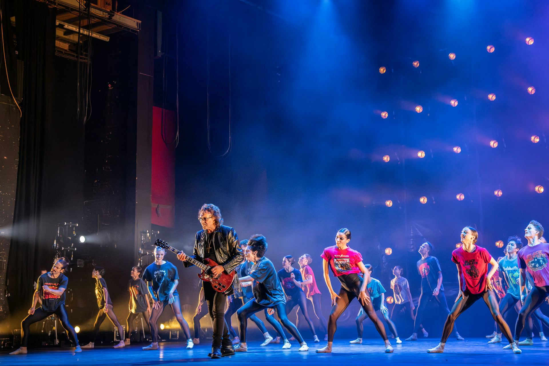 Tony I omni plays guitar on stage surrounded by dancers