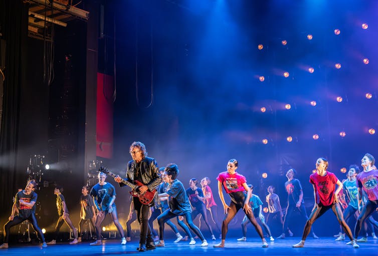 Tony I omni plays guitar on stage surrounded by dancers