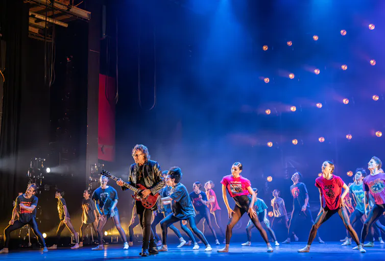 Tony I omni plays guitar on stage surrounded by dancers