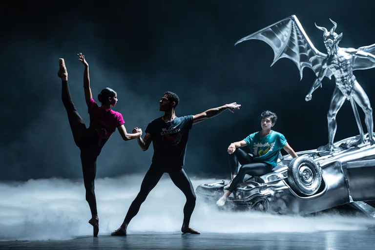 Two dancers holding hands dancing in front of a model of an upturned car.