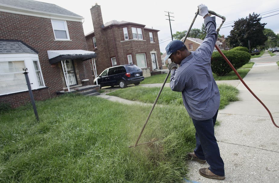 A Black man wearing a baseball cap uses a long tool in front of a modest brick home.