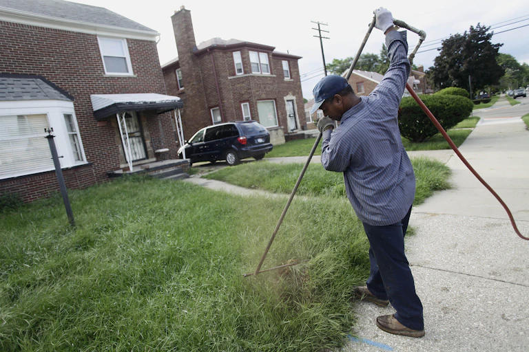 A Black man wearing a baseball cap uses a long tool in front of a modest brick home.