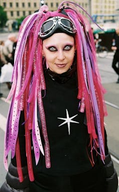 A cyber goth girl with pink hair and pale contact lenses, wearing goggles on her head.