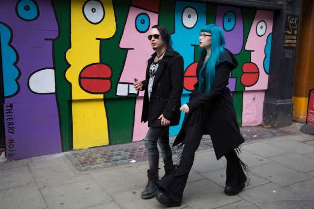 A man and woman dressed in black walk past a colourful mural. The woman has blue hair.
