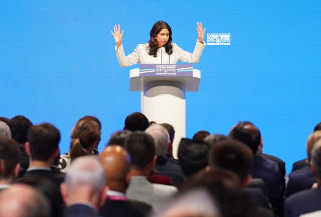 Suella Braverman with both hands raised, speaking at a podium at the Conservative party conference