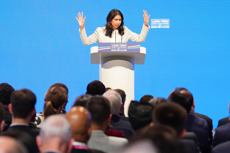 Suella Braverman with both hands raised, speaking at a podium at the Conservative party conference
