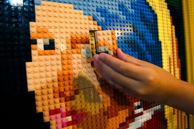 A young person's hand puts final LEGO bricks into place, finishing a sculpture of Johannes Vermeer's Girl with a Pearl Earring painting.