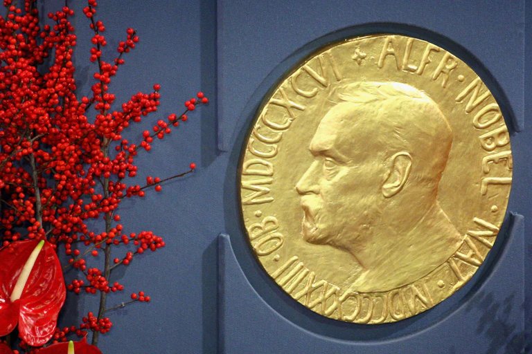 A large gold plaque of Alfred Nobel is seen against a blue wall, with red flowers nearby.