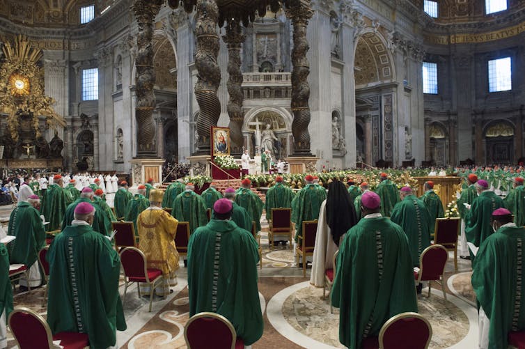 Rows of priests in green robes and pink skullcaps stand in a huge, ornate cathedral.