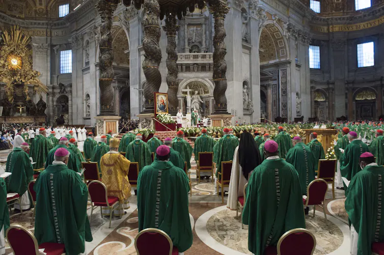 Rows of priests in green robes and pink skullcaps stand in a huge, ornate cathedral.