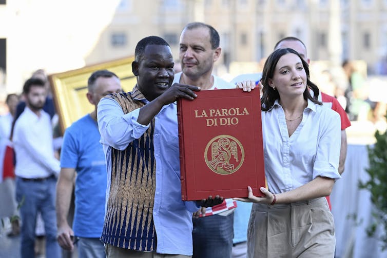 A woman and man walk holding a large red book whose cover says 'La Parola di Dio'