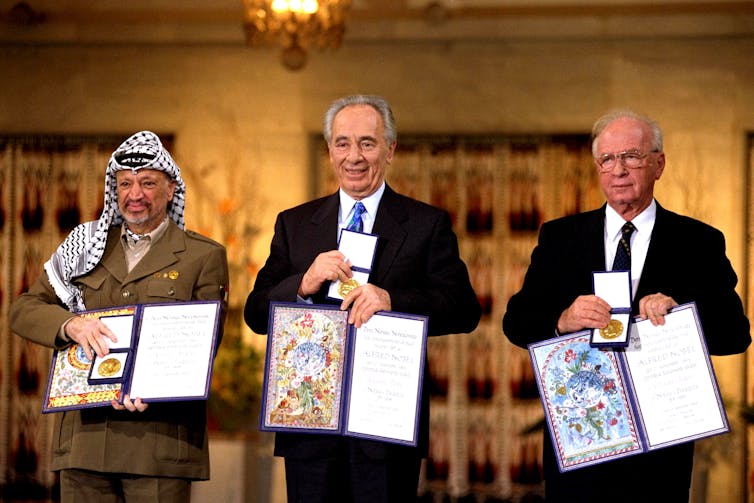 Yaser Arafat, Shimon Peres and Yitzak Rabin stand in a row and show an open book with a gold Nobel peace prize in it.