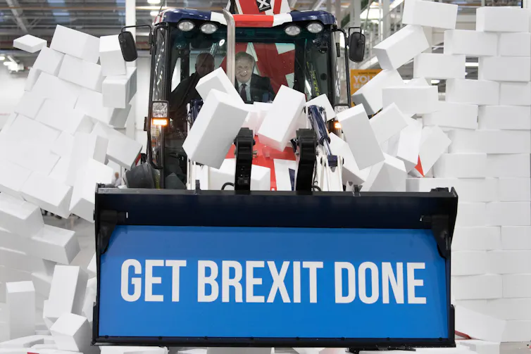 Boris Johnson driving a bulldozer with 'get Brexit done' written on its front through a wall of foam blocks.