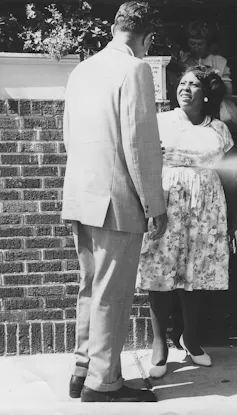 A black woman smiling and wearing a dress greets a white man in a business suit.