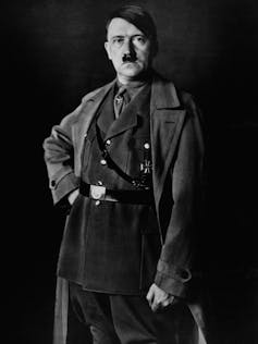 A white man with a little mustache poses while wearing a military uniform and overcoat.