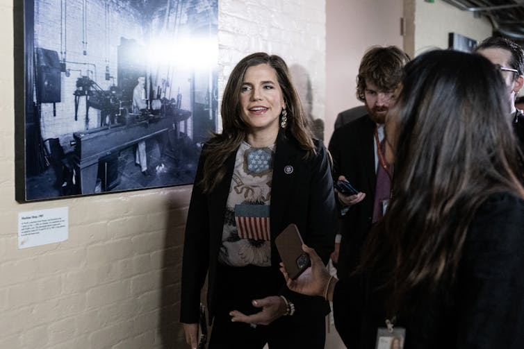 A dark-haired woman walking down a hallway, talking.