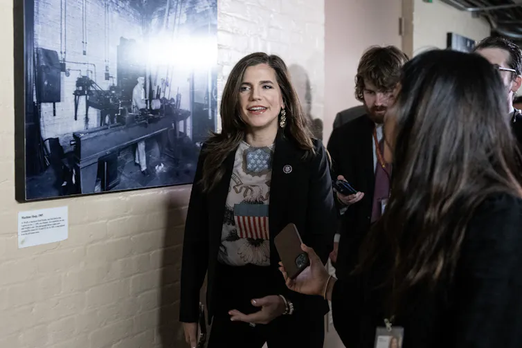 A dark-haired woman walking down a hallway, talking.
