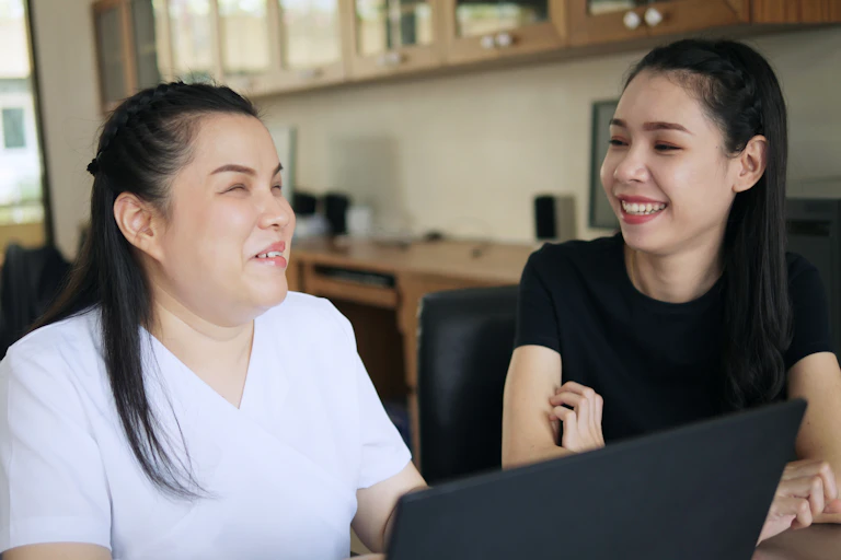 two women sit talking in front of laptop. One is vision impaired