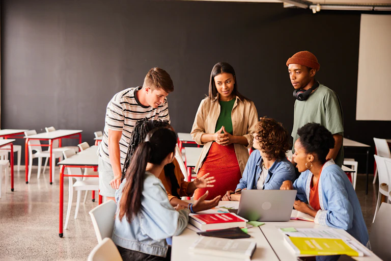 A group of seven people, some standing and some sitting, huddle together chatting. There are books and a laptop on the table in front of them