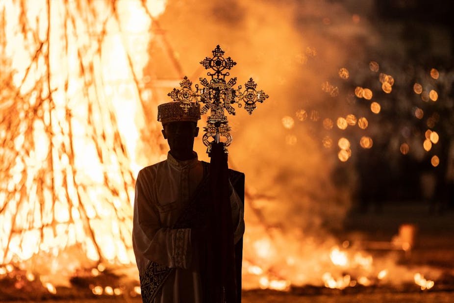 A man holding up a cross is silhouetted in front of a large fire