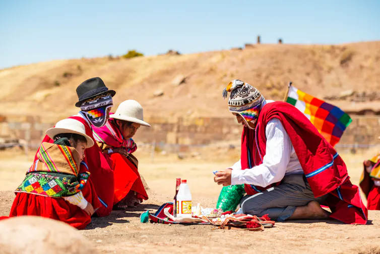 Un jeune homme autochtone aymara en vêtements traditionnels fait un rituel d'offrande à la Pachamama avec les feuilles de coca devant des enfants qui regardent.