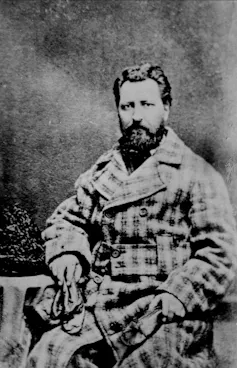 A black and white photo of a dark-haired bearded man.