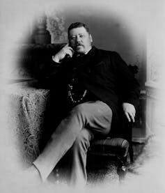A black and white photo shows a man with a moustache sitting in a chair with his legs crossed.