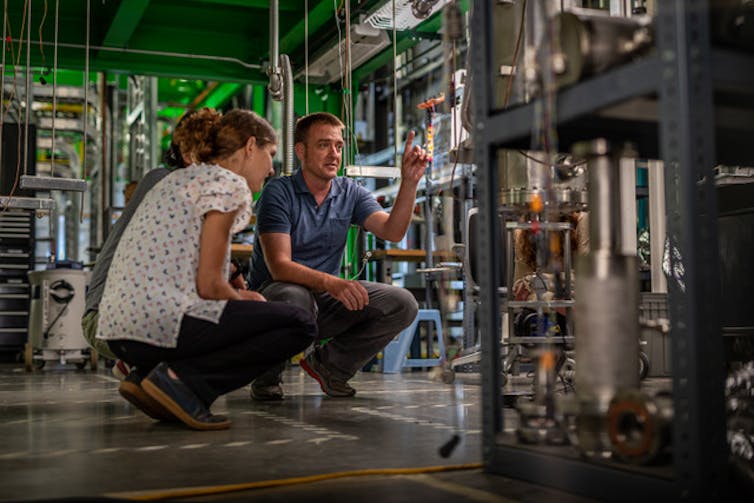 Two scientists, a woman wearing a white shirt and a man wearing a dark blue shirt, squat on the concrete ground in a laboartory with lots of machinery and shelves, and a green lit ceiling.