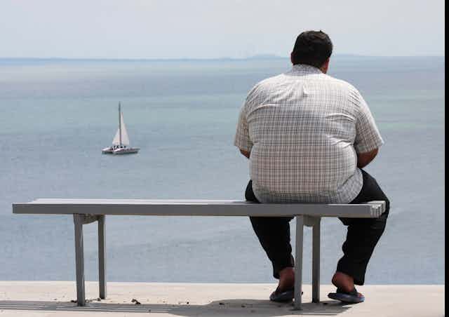 Un homme obèse est assis sur un banc, face à la mer