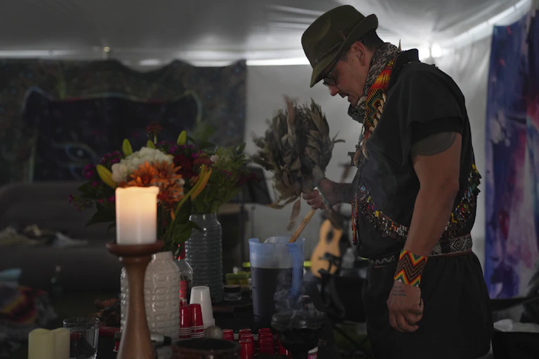 A man in a hat bowing his head before an altar on which are candles, flowers and other items.
