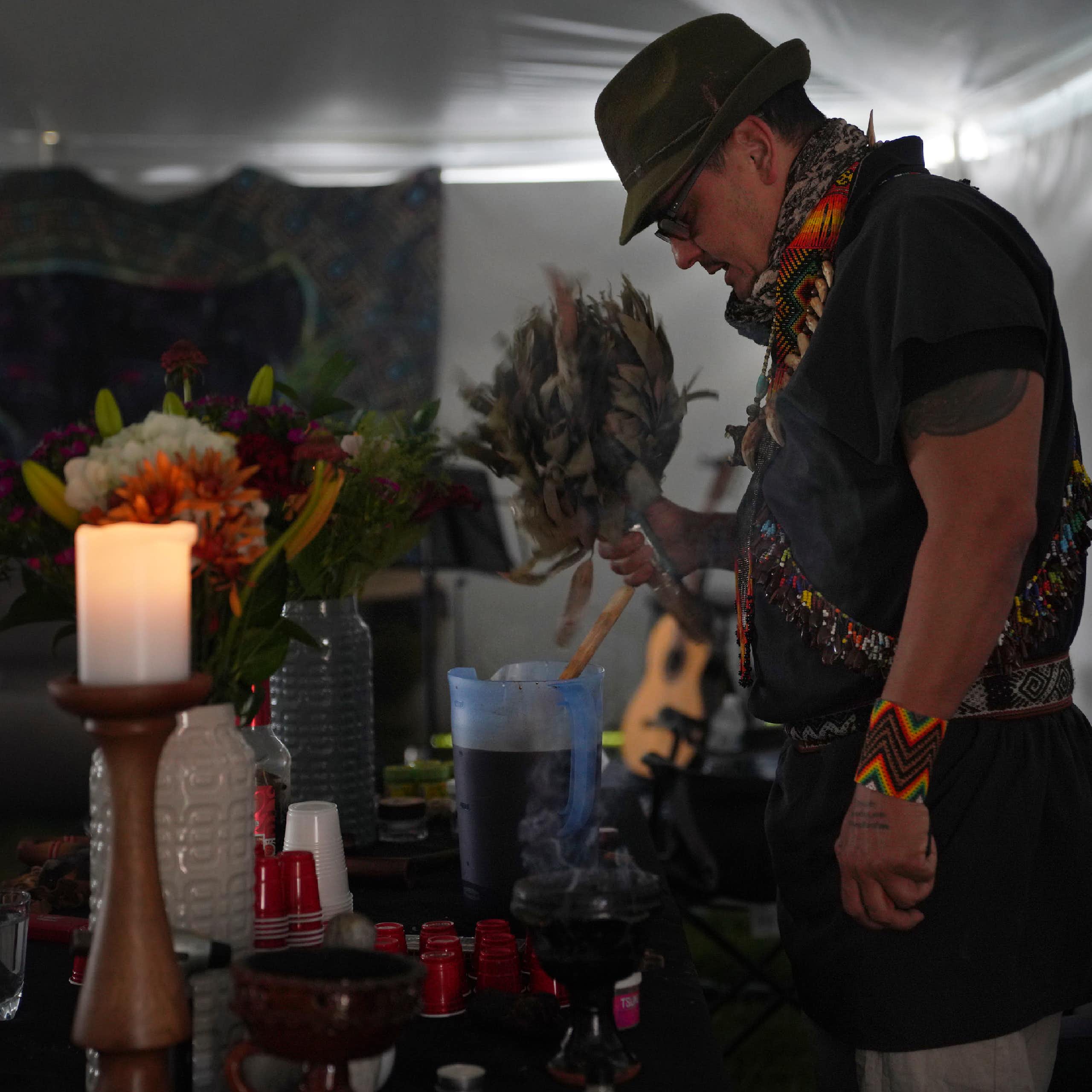 A man in a hat bowing his head before an altar on which are candles, flowers and other items.