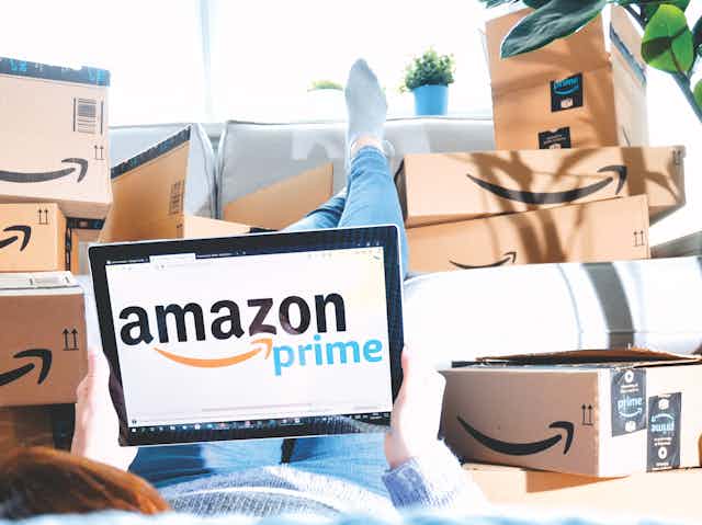 Person lying with feet up on sofa surrounded by Amazon boxes and looking at a screen with the Amazon Prime logo.
