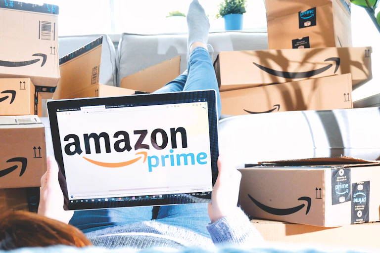 Person lying with feet up on sofa surrounded by Amazon boxes and looking at a screen with the Amazon Prime logo.