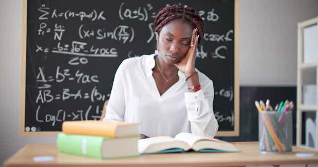 Una profesora sentada en una mesa apoya la cabeza en la mano y cierra los ojos, con una pizarra repleta de números de fondo.