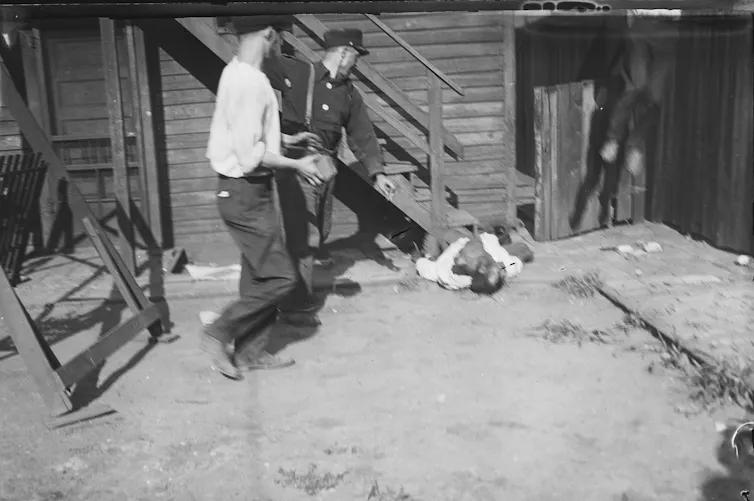 A Black man who is bleeding and laying on a sidewalk is pelted with rocks by two white men.