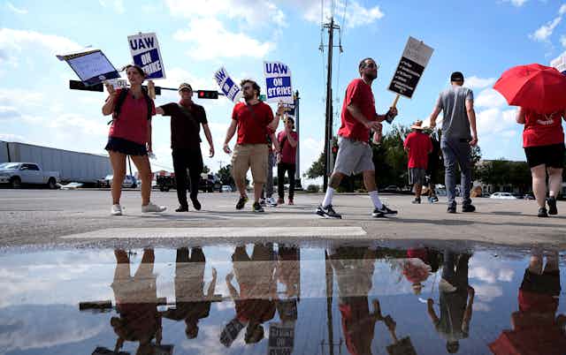 People wearing red t-shirts and carrying 'UAW on strike' placards walk in a circle near a reflective puddle.