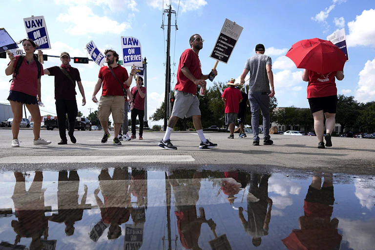 People wearing red t-shirts and carrying 'UAW on strike' placards walk in a circle near a reflective puddle.